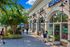 Charming retail plaza with arched storefronts, decorative lanterns, and tree-lined sidewalks as people walk, shop, and enjoy the sunny day.