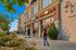 Bustling local shopping center with pedestrians walking, biking, and skating along a sidewalk lined with shops, flowers, and greenery under a clear blue sky.
