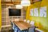 Contemporary conference room with a wooden table, ergonomic chairs, a wall-mounted TV, bold yellow accent wall, plaid wallpaper, and modern pendant lighting.