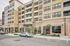 Exterior view of a modern multi-story apartment building with large windows, balconies, street-level retail space, landscaped walkways, and parked cars in front.