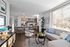 Bright and modern open-concept living space featuring a cozy sectional sofa, large window with blinds, stylish kitchen with an island and barstools, and contemporary d&eacute;cor throughout.