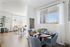 Bright apartment dining nook with a round wooden table set for four, blue upholstered chairs, large window with sheer curtains, and a view into a cozy, sunlit living room.