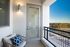 Cozy apartment balcony with a cushioned chair, wall-mounted light fixture, and railing overlooking a street and scenic landscape.