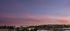 Scenic cityscape at sunset with a view of low-rise buildings, trees, rolling hills, and a sky filled with soft pink and purple clouds.
