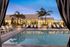 Evening view of a modern outdoor pool area with white lounge chairs, palm trees, and a lit clubhouse in the background, framed by striped cabana curtains.