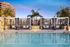Evening view of a modern poolside area featuring striped black-and-white cabanas with lounge seating, surrounded by palm trees and urban buildings.