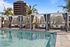 Daytime view of poolside cabanas with black-and-white striped curtains and modern lounge seating, surrounded by palm trees and urban buildings.