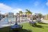 Sunny rooftop pool area with lounge chairs, striped umbrellas, palm trees, and shaded dining tables on artificial grass.