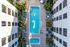 Aerial view of a rectangular outdoor swimming pool and hot tub surrounded by lounge chairs, palm trees, and patio seating, nestled between two modern apartment buildings.