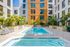 Ground-level view of a rectangular pool in a courtyard setting, surrounded by modern apartment buildings, palm trees, and neatly arranged lounge chairs.