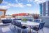 Outdoor rooftop terrace with a large dining table surrounded by blue cushioned chairs, city views in the background, and potted plants adding a touch of greenery.