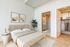A serene, modern bedroom with a light wood bed frame, neutral bedding, and minimalist decor. The room features a side table with flowers, two framed mountain prints, and direct access to a spacious walk-in closet and ensuite bathroom with a glass shower.