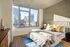 Bright bedroom with a large window offering a city view, featuring a bed with colorful pillows, modern nightstands, and a concrete accent wall.