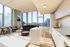 A bright, modern apartment with floor-to-ceiling windows offering expansive city views. The open-concept space includes a sleek kitchen with white cabinetry, a long island with stools, a cozy living area with a black sectional, accent chairs, and a coffee table.