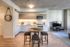A cozy open-concept kitchen and living space with modern cabinetry, marble backsplash, and a central round wooden table surrounded by three upholstered stools. A TV sits on a low black stand near the window, and a rustic hanging mirror adds charm to the entry wall.