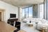 A modern, spacious living room with high ceilings, a large cream sectional sofa, marble-top coffee tables, and floor-to-ceiling windows showcasing city views. A minimalist media console and TV sit against a white wall, and the ceiling features exposed concrete for an industrial-chic touch.