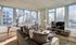 A bright, modern living room with floor-to-ceiling windows showcasing a city skyline view. The space features a plush sectional sofa, armchairs, a round coffee table, and a large TV on a sleek media stand.