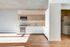 A minimalist kitchen with clean white cabinetry, integrated appliances, light wood open shelving, and natural wood flooring. Sunlight streams in, creating a warm and inviting atmosphere.