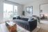 Modern living room with a dark tufted sofa, wooden coffee table, large windows letting in natural light, and a view into the bedroom.