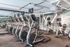 Modern gym with rows of cardio machines including ellipticals and treadmills, mirrored walls, and large windows providing natural light.