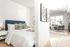 Bright studio apartment featuring a cozy bedroom area with a navy headboard, separated by a partial wall from the living and dining space with large windows.