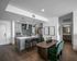 Modern open-concept apartment kitchen and dining area featuring wood flooring, light gray cabinetry, stainless steel appliances, pendant lighting, and a contemporary dining table with green velvet chairs.