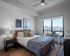 Modern bedroom with a large window overlooking the city, featuring a bed with layered pillows and throws, wooden nightstands, artwork on the wall, and a soft area rug.