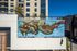 Surreal mural of a rhinoceros with tree branches, birds, and mechanical elements painted on the side of a modern building.