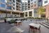 Outdoor lounge area with tables, chairs, greenery, and a life-sized chess set surrounded by modern apartment buildings.