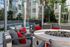 Outdoor lounge area with cushioned seating, red accent pillows, and a round fire pit centerpiece, surrounded by glass walls and greenery.