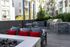 Outdoor patio area featuring a built-in stainless steel grill, prep counter, cushioned lounge seating with red pillows, and surrounding modern apartment buildings and palm trees.