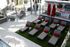 Outdoor sun deck featuring a row of cushioned lounge chairs with red accent pillows on artificial grass, surrounded by patio seating, umbrellas, and modern apartment amenities.