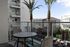 Outdoor balcony or patio area featuring a round table with chairs, palm trees, outdoor seating, and a view of nearby buildings under a clear blue sky.