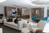 Spacious entertainment lounge with a sleek kitchen and bar seating, a long dining table with teal chairs, modern light fixtures, a mounted TV, and contemporary furniture including a cream sofa and lounge chairs.