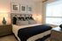 Modern bedroom with a neatly made bed featuring neutral-toned bedding and navy accents, two matching nightstands with lamps, framed artwork above the headboard, and a large window letting in natural light.