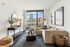 A modern living room with a beige sectional sofa, round coffee table, city-view window, console table with d&eacute;cor, framed artwork, and a large mirror, all illuminated by natural light on dark wood flooring.