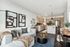 A stylish open-layout living room with a cushioned sofa, abstract wall art, a decorative coffee table, and a cozy kitchen in the background featuring white cabinets, stainless steel appliances, and a kitchen island with bar stools.