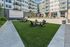 Outdoor courtyard at Brickline apartment complex featuring Adirondack chairs around fire pits on a green lawn, surrounded by modern apartment buildings with balconies and red patio umbrellas.