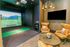 Indoor golf simulator room with a virtual driving range, cushioned chairs, a round table, and a wall-mounted TV.