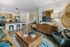 Living room with modern furnishings, a wooden media console, colorful decor, and a view into a kitchen with a breakfast bar and pendant lighting.