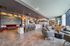 A spacious, modern resident lounge with a large sectional sofa, armchairs, high ceilings with exposed beams, polished concrete flooring, and oversized windows bringing in natural light, with a kitchen and fireplace enhancing the open, cozy atmosphere.