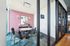 A modern coworking space featuring a glass-walled meeting room with a pink accent wall, a rectangular table surrounded by chairs, and a wall-mounted monitor. The room is set on a blue and gray patterned carpet, with views into an adjacent area containing shelving and office furnishings.