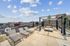 An outdoor rooftop patio featuring lounge chairs, a seating area with cushioned armchairs surrounding a fire pit, and a pergola overhead. The space is enclosed by a railing and offers cityscape views, with a partly cloudy sky adding to the relaxed, social atmosphere.