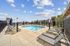 An outdoor pool area featuring several lounge chairs with umbrellas arranged around a rectangular swimming pool, bordered by a safety fence. Surrounding buildings and a clear sky with scattered clouds frame the relaxing scene, offering a tranquil, resort-style ambiance.