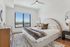 Modern bedroom featuring a curved headboard, layered bedding, large windows with city views, a ceiling fan, and a neutral-toned rug.