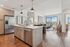 Modern open kitchen with a large island, stainless steel appliances, pendant lighting, and a view into a sunlit living area with large windows.