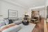 Bright and airy living room with a large light-gray sectional sofa, minimalist wall art, round coffee table, and open layout connecting to a dining and kitchen area with wood cabinetry.