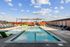 A modern rooftop pool deck with lounge chairs, green poolside loungers, and a shaded pergola seating area, set under a bright blue sky with scattered clouds.