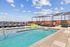 Bright and modern rooftop pool area featuring green lounge chairs in a shallow section, deeper swimming pool, shaded pergola seating, and skyline views under a blue sky.