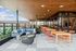 Modern rooftop space featuring outdoor dining tables, lounge seating, string lighting, and a wooden ceiling, overlooking the city skyline.